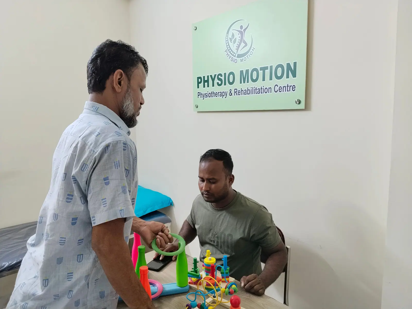 A physiotherapist at Physio Motion guiding a seated patient through hand and finger coordination exercises using coloured therapy rings, beneath the clinic’s wall sign in Uttara