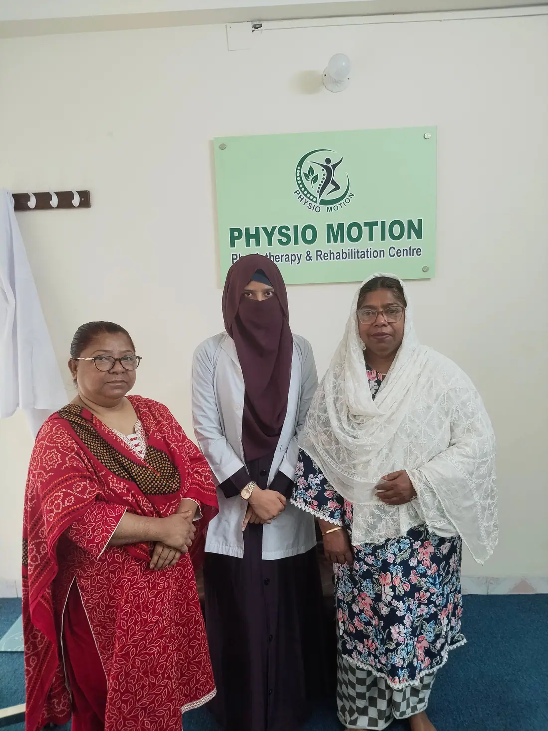 A female physiotherapist in a white coat standing with two women patients beneath the Physio Motion sign at the Uttara centre