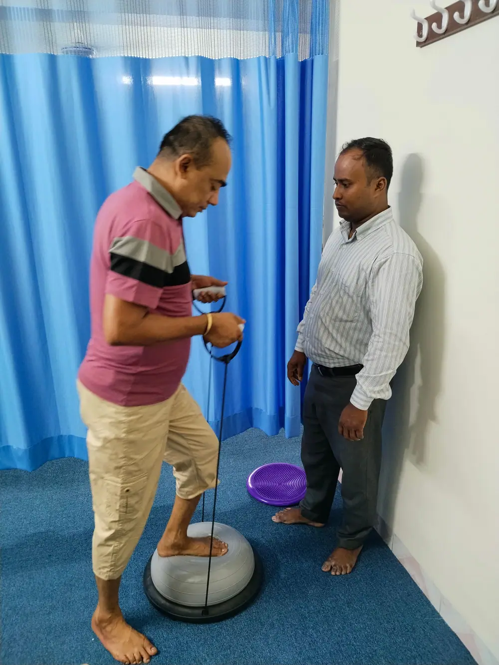 A patient balancing on a wobble trainer while pulling a resistance band, supervised by a physiotherapist at Physio Motion